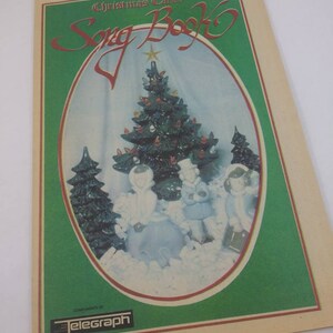 May include: A vintage Christmas Carol Song Book with a green border and a cream-colored background. The cover features a Christmas tree and figures in a snowy scene. The text "Christmas Carol Song Book" is in red script. The Telegraph logo is at the bottom.