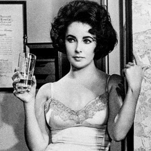 Puede incluir: Una fotografía en blanco y negro de una mujer con un vestido blanco de encaje. Ella está sosteniendo un vaso de agua y mirando hacia un lado.