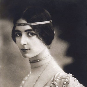 Pode incluir: Um retrato em preto e branco de uma mulher usando uma faixa de cabeça com contas e um vestido com detalhes de pérolas. O texto "REUTLINGER PARIS." é visível no canto inferior esquerdo da imagem.