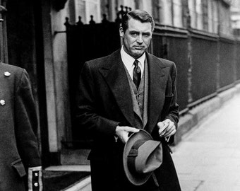 May include: A man in a suit and overcoat walks down a street, holding a fedora in his hand. He is wearing a tie and a vest. The photo is in black and white.