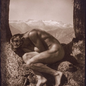 Puede incluir: Una figura desnuda se sienta de espaldas a la cámara, encorvada con la cabeza apoyada en las rodillas. La figura está sentada en un tronco de árbol con una cordillera de montañas al fondo.