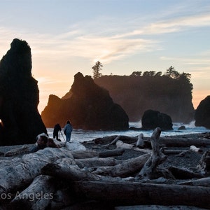 Op de afbeelding: Silhouetten van mensen die op een rotsachtig strand staan ​​bij zonsondergang. De lucht is fel oranje en roze, en het water is diepblauw. De rotsen zijn donker en grillig, en het strand is bedekt met drijfhout.