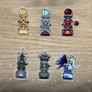 May include: Six clear acrylic charms with colorful designs. The charms feature a pillar-like shape with various colors, including gold, red, blue, and silver. Each charm has a unique design, with geometric and fantasy-inspired elements.