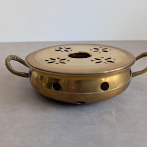 Puede incluir: Un calentador redondo de latón con dos asas laterales curvas y una tapa extraíble con cuatro patrones florales recortados y una abertura central circular. La base del calentador tiene una serie de pequeños orificios de ventilación circulares.