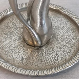 May include: A silver ring holder in the shape of a cat with a decorative, embossed tray. The tray has a floral pattern.
