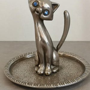 May include: Silver cat ring holder with blue rhinestone eyes, sitting on a decorative tray.