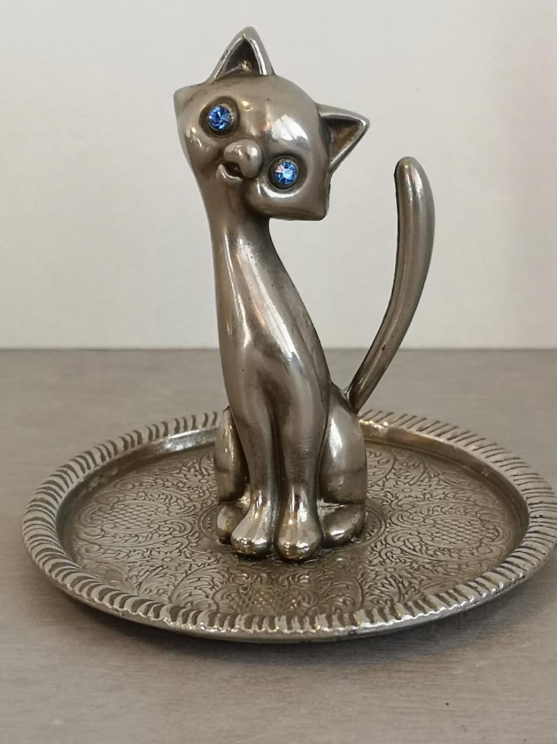 May include: Silver cat ring holder with blue rhinestone eyes, sitting on a decorative tray.