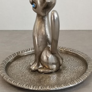 May include: Silver cat ring holder with blue rhinestone eyes. The cat is sitting on a small, round, silver tray with an ornate design.