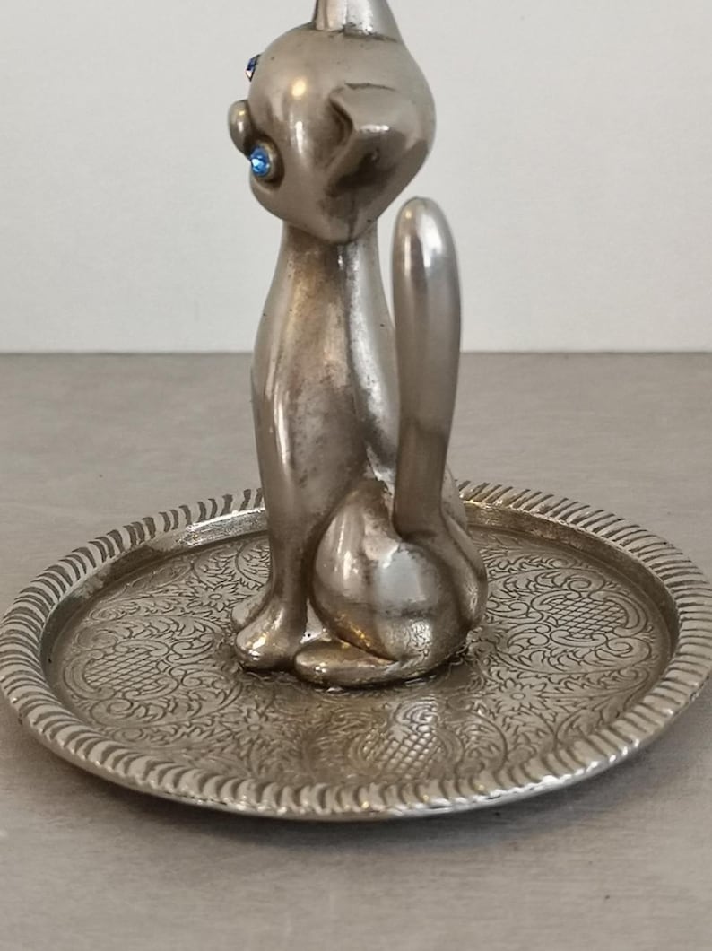 May include: Silver cat ring holder with blue rhinestone eyes. The cat is sitting on a small, round, silver tray with an ornate design.