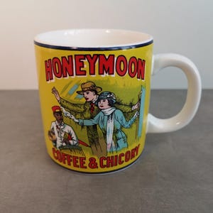 Op de afbeelding: Een witte keramische mok met een gele achtergrond en een vintage illustratie van een stel op huwelijksreis. De tekst "Honeymoon Coffee & Chicory" staat op de mok gedrukt.