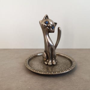 May include: Silver ring holder shaped like a cat with blue eyes. The cat is sitting on a small, round, silver tray with an ornate design.