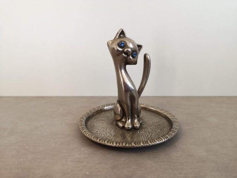 May include: Silver ring holder shaped like a cat with blue eyes. The cat is sitting on a small, round, silver tray with an ornate design.