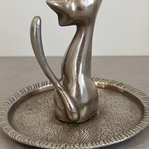May include: Silver ring holder shaped like a cat sitting on a decorative tray. The tray has an intricate floral design.