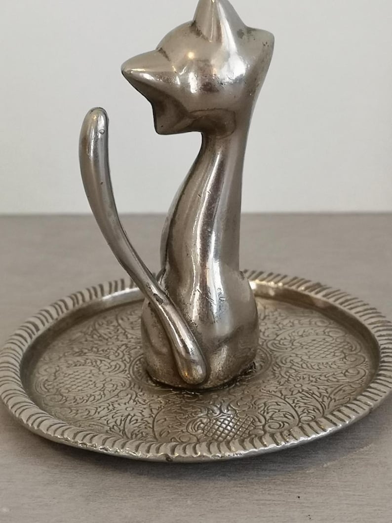 May include: Silver ring holder shaped like a cat sitting on a decorative tray. The tray has an intricate floral design.