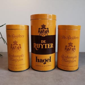 Op de afbeelding: Drie vintage gele metalen blikken met bruine etiketten. De blikken zijn gelabeld "De Ruyters" en hebben een wapenschild met een kroon. De blikken zijn gelabeld "Gestampte Muisjes", "Hagel" en "Gestampte Muisjes".