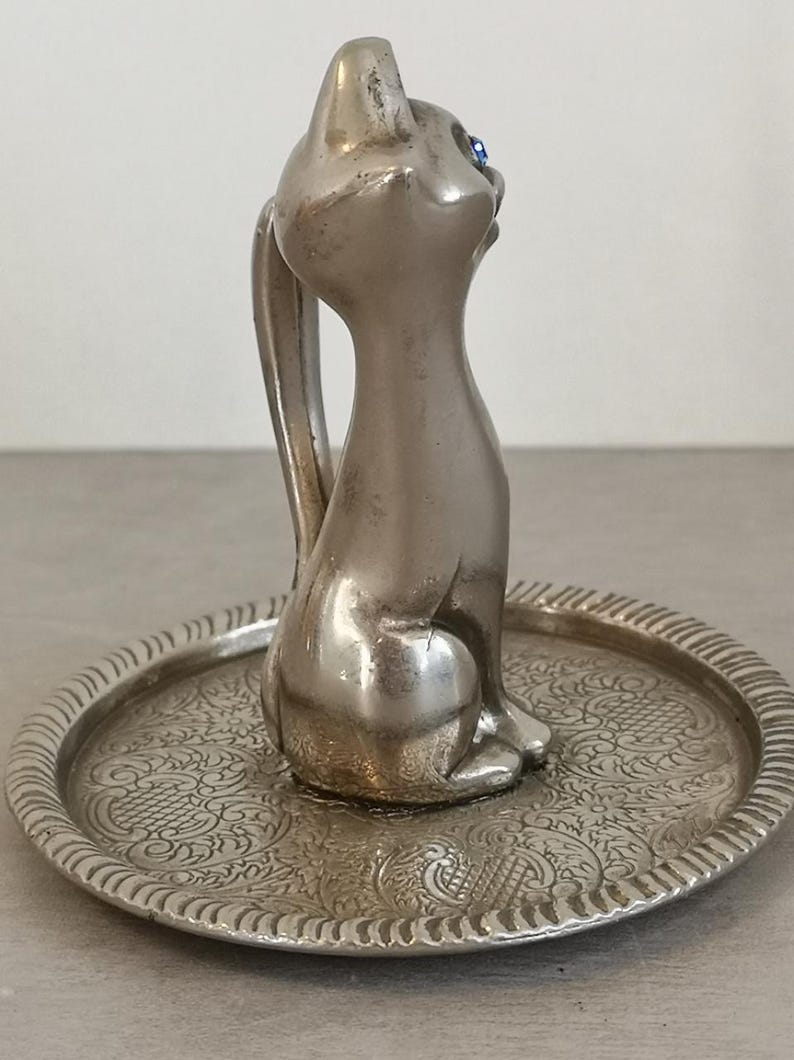 May include: Silver ring holder featuring a cat figurine with blue eyes sitting on a decorative tray.
