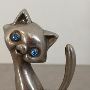 May include: A silver-colored cat figurine with blue rhinestone eyes. The cat is sitting with its tail curled around its body.