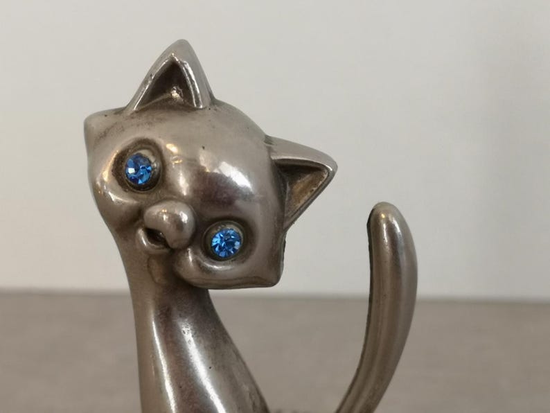 May include: A silver-colored cat figurine with blue rhinestone eyes. The cat is sitting with its tail curled around its body.