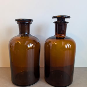 Op de afbeelding: Twee amberkleurige glazen apothekersflessen met brede, ronde lichamen en korte, smalle halzen. Elke fles is voorzien van een platte, ronde glazen stop die in de opening rust. Het glas is doorschijnend en glad en reflecteert het licht op het oppervlak.