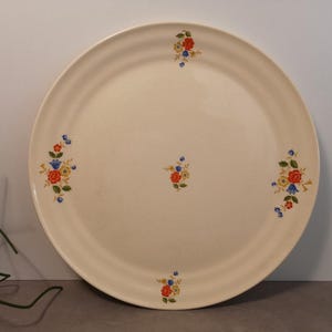 Może przedstawiać: Biały talerz ceramiczny z kwiatowym wzorem. Talerz ma podwyższony rant i jest ozdobiony małymi skupiskami czerwonych, żółtych i niebieskich kwiatów.