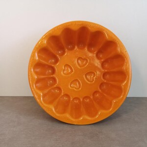 Op de afbeelding: Een ronde, oranje keramische bakvorm met een gegolfde rand en hartvormige details. De vorm is bedoeld om te bakken en staat op een grijze ondergrond. Diameter ongeveer 20 cm.
