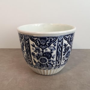 Op de afbeelding: Een witte keramische kom met een blauw bloemmotief. De kom heeft een afgeronde vorm met een licht uitlopende rand. Het blauwe patroon bevat bloemen en andere decoratieve elementen. De basis van de kom heeft een geribbeld ontwerp.