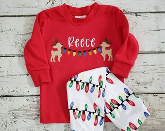 May include: Red long-sleeved pajama top and pants set. The top features the name "Reece" in white script, with reindeer and a string of colorful Christmas lights. The pajama pants are white with a repeating pattern of Christmas lights.
