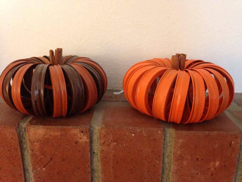 May include: Two pumpkins made from stacked orange and brown jar lids. The pumpkins are sitting on a brick surface.