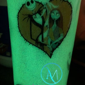 Puede incluir: Un vaso verde que brilla en la oscuridad con un dise&ntilde;o de coraz&oacute;n cosido con Jack Skellington y Sally de Pesadilla antes de Navidad. El vaso tambi&eacute;n tiene el texto "AmMommie Designs" en la parte inferior.