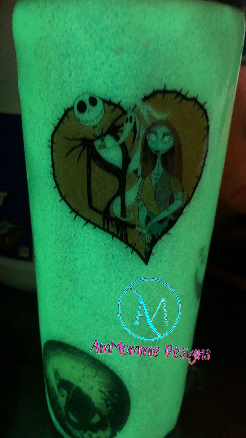Puede incluir: Un vaso verde que brilla en la oscuridad con un dise&ntilde;o de coraz&oacute;n cosido con Jack Skellington y Sally de Pesadilla antes de Navidad. El vaso tambi&eacute;n tiene el texto "AmMommie Designs" en la parte inferior.