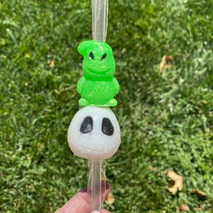 May include: A green and white character-themed straw with a green character with a toothy grin on top of a white character with black eyes. The straw is clear plastic.