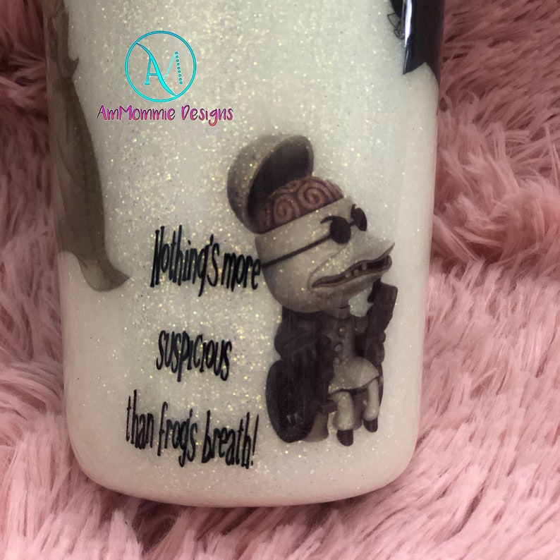 Puede incluir: Un vaso blanco con purpurina con una ilustraci&oacute;n en blanco y negro de un personaje de la pel&iacute;cula Pesadilla antes de Navidad. El personaje lleva un sombrero y gafas de sol. El texto "Nothing's more suspicious than frogs breath!" est&aacute; impreso en el vaso.