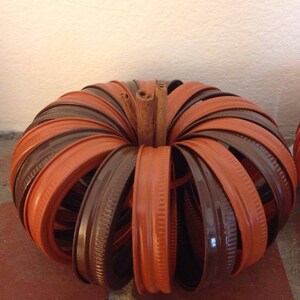 May include: A pumpkin-shaped craft made from brown and orange jar lids. The lids are arranged in a circular pattern with a cinnamon stick in the centre.