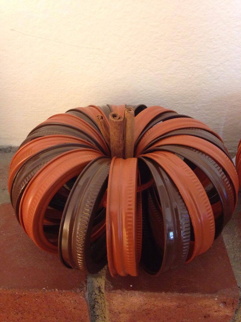 May include: A pumpkin-shaped craft made from brown and orange jar lids. The lids are arranged in a circular pattern with a cinnamon stick in the centre.