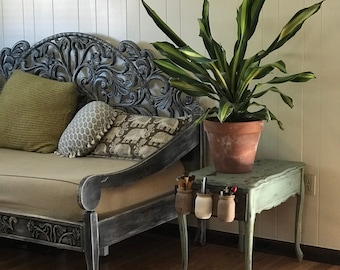 May include: A light green side table with four cabriole legs and three small mason jars attached to the front edge, holding various pens and brushes. A large potted plant with green and yellow variegated leaves sits on the table surface.