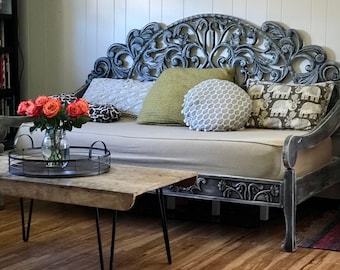 May include: A daybed with a distressed gray carved wooden frame and a beige cushion, featuring several decorative pillows in green, white, and elephant patterns. A wooden coffee table with black metal hairpin legs sits in front.