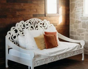 May include: A white daybed featuring an ornate, carved wooden backrest with intricate scrollwork and floral motifs. The frame includes matching carved detailing along the front rail. The bed is dressed with white linens and three decorative throw pillows.