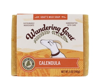May include: A bar of goat's milk soap with a yellow and orange label. The label features a cartoon goat and the text "Wandering Goat" and "Calendula". The soap is made with sustainable ingredients and handcrafted with care. It weighs 140 grams.