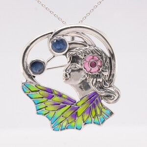 May include: Silver pendant necklace featuring a woman's face with a pink flower in her hair, surrounded by a circle with two blue gemstones. The pendant is attached to a silver chain and has a butterfly design with green, blue, and purple enamel.
