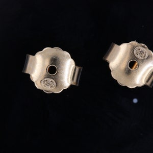 May include: Close-up of two gold-toned earring backs. Each back has a small hole in the center and a scalloped edge. The backs are marked with the letters "14K".