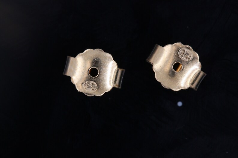 May include: Close-up of two gold-toned earring backs. Each back has a small hole in the center and a scalloped edge. The backs are marked with the letters "14K".