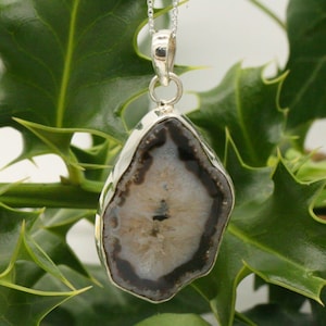 Puede incluir: Un colgante de plata con una rebanada de ágata natural con un patrón gris y negro. El ágata está engastada en un marco de plata y cuelga de una cadena de plata.