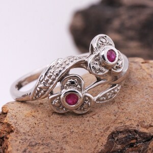 May include: A silver ring with a floral design. The ring features two flower clusters, each with a round, red gemstone centre. The ring has a textured band and is set on a natural, brown surface.