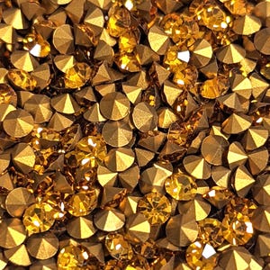 Puede incluir: Una imagen de primer plano de una gran cantidad de pequeños diamantes de imitación redondos de color dorado. Los diamantes de imitación tienen un diseño facetado en forma de pirámide.