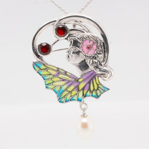 Puede incluir: Un colgante de plata con un diseño de mariposa de esmalte colorido, una cara de mujer y piedras preciosas rojas y rosas. Una perla cuelga de la parte inferior.