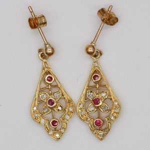 Puede incluir: Un par de pendientes de oro con un diseño delicado e intrincado. Los pendientes tienen forma de lágrima con un motivo floral y están adornados con pequeñas piedras preciosas rojas y blancas.