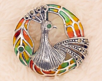Puede incluir: Un broche de pavo real de plata con detalles de esmalte verde y naranja y marcasita. El pavo real se representa con las plumas de la cola extendidas.