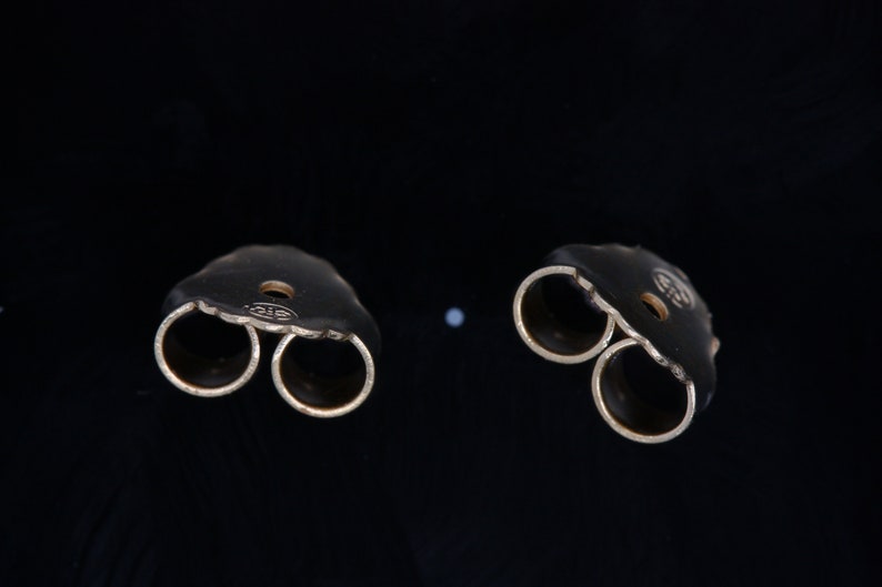 May include: Two gold-toned earring backs with a double-prong design. The backs are designed to hold a stud earring in place.