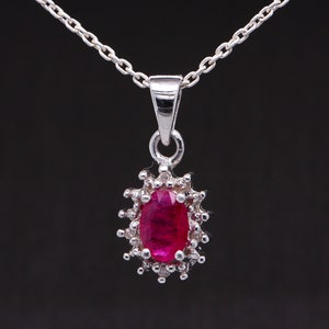 May include: A silver necklace with a pendant featuring a red oval gemstone surrounded by smaller clear gemstones.