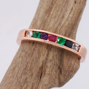 Puede incluir: Anillo de oro rosa con una fila de piedras preciosas de colores. El anillo presenta piedras blancas, verdes, moradas, rojas, negras y transparentes, engastadas en un canal. El anillo se muestra sobre un trozo de madera.